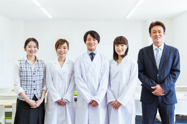 中央に白衣を着て笑顔で立つ医療従事者３人と両端に笑顔で立つ事務員２人のスナップ写真